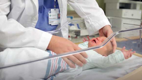 A newborn baby lies in a hospital bassinet cart while being examined by a family medicine resident.